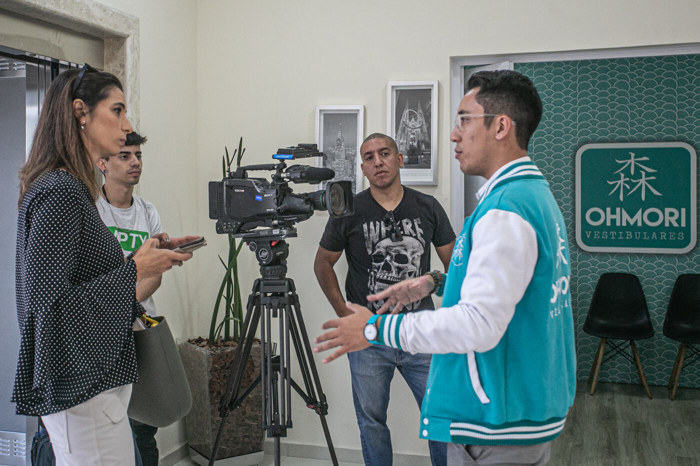 Marco, de jaqueta Ohmori, dá entrevista à repórter da Globo diante da câmera, na recepção da escola