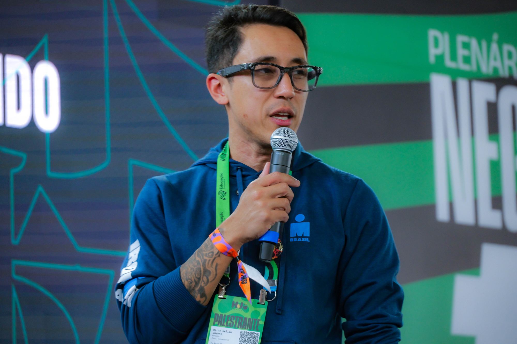 Marco Ohmori de óculos, microfone na mão e crachá de palestrante, na Plenária Negócios 2026